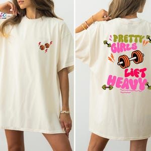 Puede incluir: Camiseta color crema con un peque&ntilde;o gr&aacute;fico de mancuernas y un rayo en la parte delantera. La parte trasera presenta el texto "Pretty Girls Lift Heavy" con un gr&aacute;fico de mancuernas. El dise&ntilde;o utiliza colores naranja, verde y rosa.