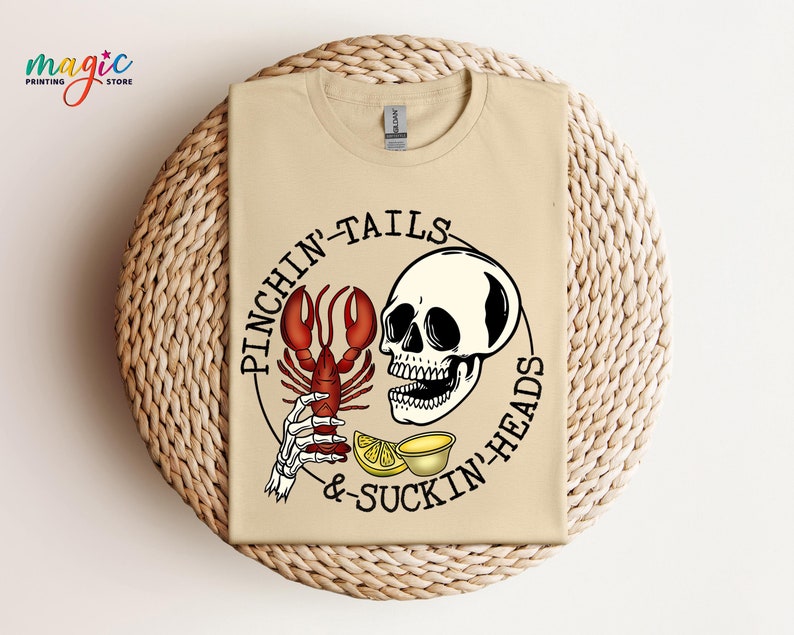 K&ouml;nnte beinhalten: Ein beiges T-Shirt mit einer Schwarzwei&szlig;illustration eines Sch&auml;dels, der eine Hummerklaue h&auml;lt. Der Text "Pinchin' Tails & Suckin' Heads" ist in einem Kreis um den Sch&auml;del geschrieben.