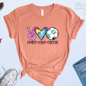 Może przedstawiać: Koszulka w kolorze brzoskwiniowym z grafiką przedstawiającą znak pokoju, serce i piłkę nożną. Grafika jest w stylu tie-dye w kolorach r&oacute;żowym, niebieskim i ż&oacute;łtym. Pod grafiką znajduje się napis "Peace Love Soccer".