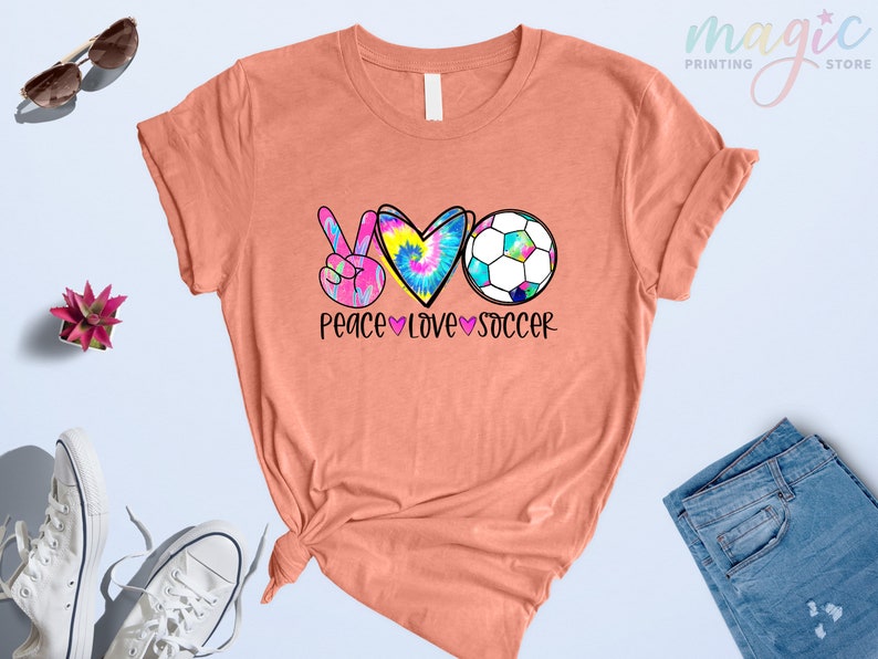 Może przedstawiać: Koszulka w kolorze brzoskwiniowym z grafiką przedstawiającą znak pokoju, serce i piłkę nożną. Grafika jest w stylu tie-dye w kolorach r&oacute;żowym, niebieskim i ż&oacute;łtym. Pod grafiką znajduje się napis "Peace Love Soccer".