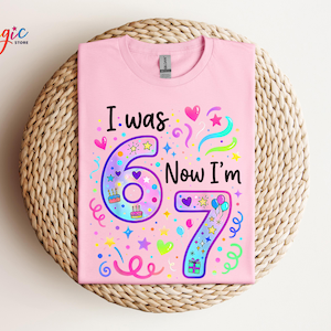 May include: A pale pink t-shirt featuring the text "I was 6 Now I'm 7" in black and colourful graphics. The number 6 is purple and the number 7 is blue. The shirt has a crew neck.