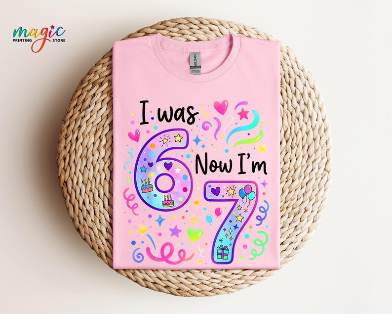 May include: A pale pink t-shirt featuring the text "I was 6 Now I'm 7" in black and colourful graphics. The number 6 is purple and the number 7 is blue. The shirt has a crew neck.