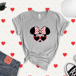 May include: A gray t-shirt with a black and white illustration of Minnie Mouse wearing red heart-shaped sunglasses and a red and white polka dot bow. The background is a white pattern of red hearts.