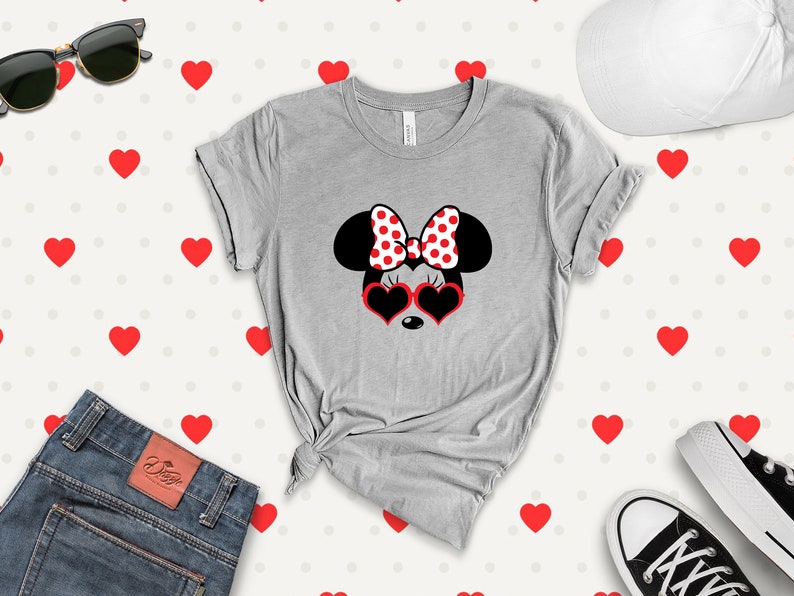 May include: A gray t-shirt with a black and white illustration of Minnie Mouse wearing red heart-shaped sunglasses and a red and white polka dot bow. The background is a white pattern of red hearts.