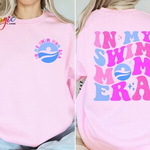 May include: Pink sweatshirt with the text "In My Swim Mom Era" in a retro font with blue, pink, and white colours. The text is on the back of the sweatshirt. The front of the sweatshirt has a smaller version of the text in a circle.
