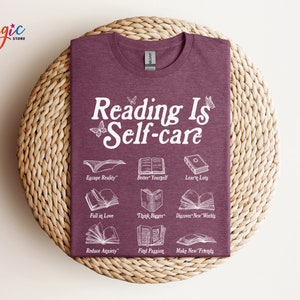 May include: A maroon t-shirt with the text "Reading Is Self-care" in white. The shirt features illustrations of open books with text describing the benefits of reading, such as "Escape Reality", "Better Yourself", "Learn Lots", "Fall in Love", "Think Bigger", "Discover New Worlds", "Reduce Anxiety", "Find Passion", and "Make New Friends".