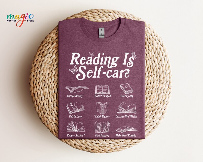 May include: A maroon t-shirt with the text "Reading Is Self-care" in white. The shirt features illustrations of open books with text describing the benefits of reading, such as "Escape Reality", "Better Yourself", "Learn Lots", "Fall in Love", "Think Bigger", "Discover New Worlds", "Reduce Anxiety", "Find Passion", and "Make New Friends".