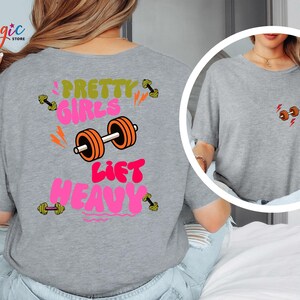 Puede incluir: Camiseta gris con el texto "Pretty Girls Lift Heavy" en rosa, verde y naranja. El dise&ntilde;o incluye ilustraciones de pesas. Una versi&oacute;n m&aacute;s peque&ntilde;a del gr&aacute;fico de la pesa est&aacute; en la parte delantera.