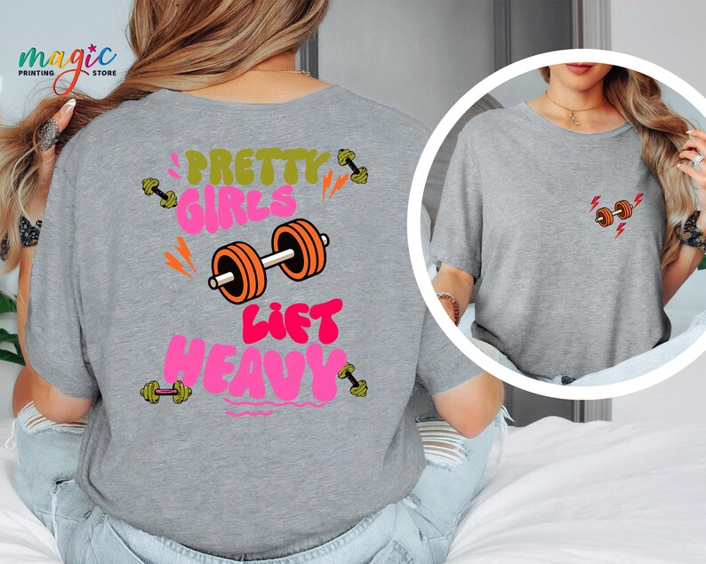Puede incluir: Camiseta gris con el texto "Pretty Girls Lift Heavy" en rosa, verde y naranja. El dise&ntilde;o incluye ilustraciones de pesas. Una versi&oacute;n m&aacute;s peque&ntilde;a del gr&aacute;fico de la pesa est&aacute; en la parte delantera.