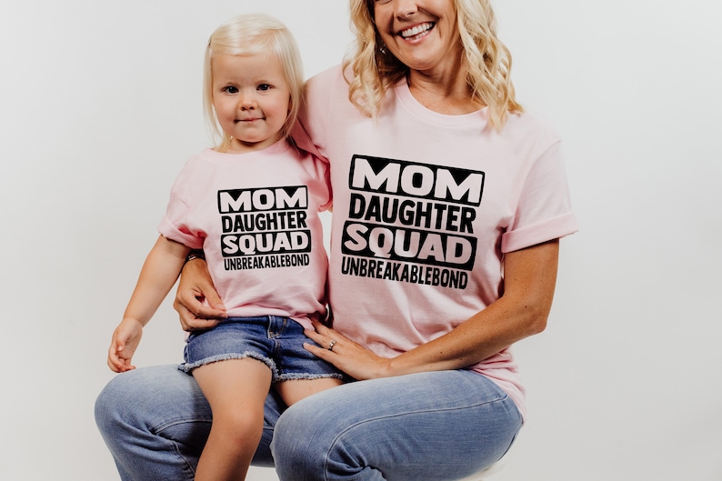 May include: A pink t-shirt with the text "Mom Daughter Squad Unbreakablebond" in black. A child is wearing a smaller version of the same shirt.
