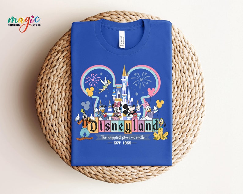Puede incluir: Camiseta azul real con un gr&aacute;fico colorido de Disneyland. El dise&ntilde;o incluye el logotipo de Disneyland, un castillo, fuegos artificiales y varios personajes de Disney. El texto dice "Disneyland, The happiest place on earth, EST. 1955."