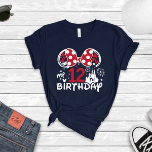 Pu&ograve; includere: Una maglietta blu navy con una grafica bianca di un fiocco rosso e bianco a pois, il numero 12, un castello e fuochi d'artificio. Il testo "my 12th birthday" &egrave; stampato sotto la grafica.