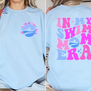 May include: A light blue sweatshirt with the text "In My Swim Mom Era" in pink and blue letters. The text is in a retro font style. The sweatshirt has a small blue circle with a swimmer icon on the left side of the chest.