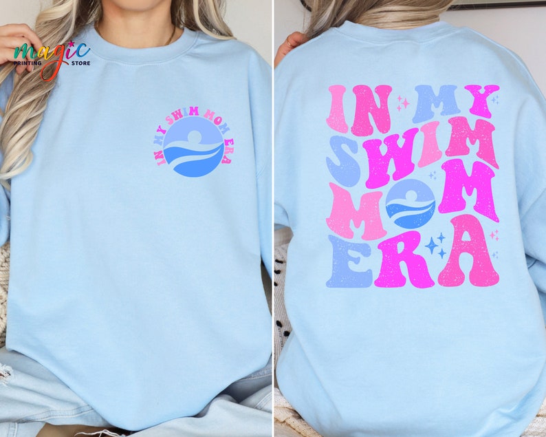 May include: A light blue sweatshirt with the text "In My Swim Mom Era" in pink and blue letters. The text is in a retro font style. The sweatshirt has a small blue circle with a swimmer icon on the left side of the chest.