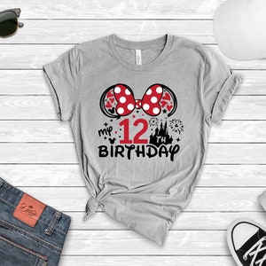 Pu&ograve; includere: Maglietta grigia con un fiocco di Minnie Mouse rosso e bianco, un castello, fuochi d'artificio e il testo "my 12th birthday".