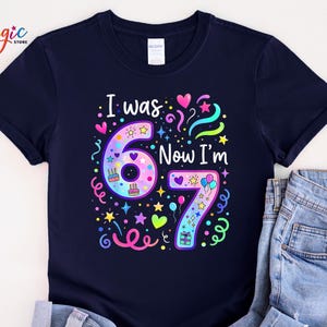 May include: A navy blue t-shirt featuring the text "I was 67 Now I'm 67" in a vibrant design. The design incorporates the numbers 6 and 7, hearts, stars, balloons, and confetti, ideal for a birthday celebration.