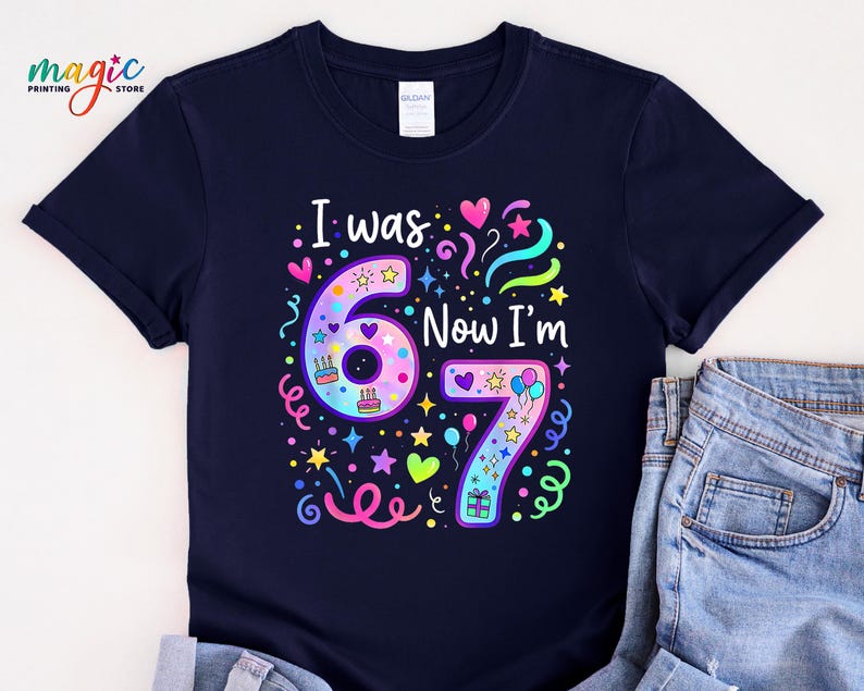 May include: A navy blue t-shirt featuring the text "I was 67 Now I'm 67" in a vibrant design. The design incorporates the numbers 6 and 7, hearts, stars, balloons, and confetti, ideal for a birthday celebration.