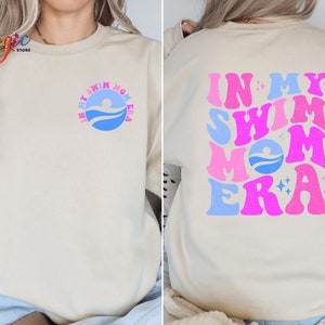 May include: A cream-coloured sweatshirt with the text "In My Swim Mom Era" in pink, blue and white letters. The text is in a retro font style with stars and a swimmer graphic.