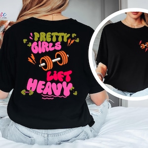 Puede incluir: Camiseta negra con el texto "PRETTY GIRLS LIFT HEAVY" en rosa y verde, con un gr&aacute;fico de barra con pesas. Un gr&aacute;fico de barra con pesas m&aacute;s peque&ntilde;o est&aacute; en la parte delantera. La camiseta es de estilo casual.
