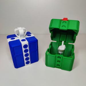 May include: Two 3D-printed boxes. One is blue and white, resembling a gift box with a bow. The other is green and open, revealing a white hand giving the middle finger. The boxes are on a white surface.