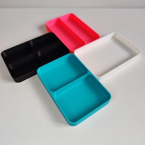 May include: Four brightly coloured plastic trays in black, pink, white, and teal. The black tray has five compartments, the pink tray has two, the white tray has one, and the teal tray has two. These trays are ideal for organising small items.