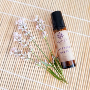 Puede incluir: Un frasco de vidrio marrón con tapa negra y una etiqueta que dice "Yantra Aromatics Lavender Hygge 10ml". El frasco está sobre una estera de bambú con una rama de flores de lavanda.