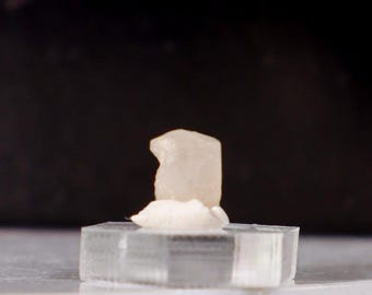 May include: A small, light-colored crystal formation atop a clear, hexagonal base. The crystal sits on a white, textured surface, with a dark background. The base is on a white marble surface with a gold trim.