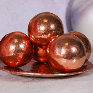 May include: A collection of four smooth, spherical objects with a reflective copper-toned metallic finish, arranged in a cluster on a matching circular, shallow, hammered copper-toned metal dish with a textured, uneven rim.