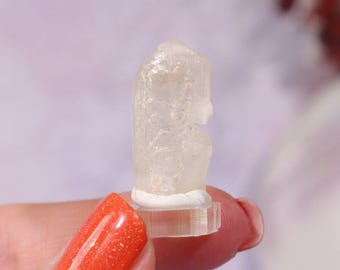 May include: A small, clear crystal figurine, possibly quartz, is held between fingers. The crystal is mounted on a clear base with a white adhesive. The figurine has a rounded shape with a small, carved detail. The person holding the crystal has orange glitter nail polish.