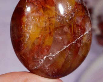 Puede incluir: Una piedra preciosa pulida y redonda con una mezcla de tonos naranja, rojo y amarillo. La piedra tiene una calidad translúcida con patrones internos y una fisura blanca visible. La gema es sostenida por una mano con esmalte de uñas azul.