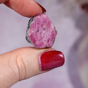 Peut inclure: Une pierre précieuse de rubis rose brute avec des bords gris foncé. Le rubis est tenu entre deux doigts, mettant en valeur sa texture naturelle non polie et sa couleur vibrante. L'arrière-plan est un violet doux et flou.