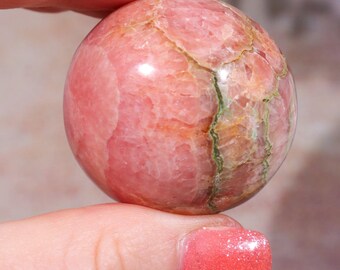 May include: A polished, spherical gemstone, primarily pink with veins of green and brown. The sphere is held between fingers, showcasing its smooth, rounded form and natural color variations. The gemstone's surface has a marbled appearance.