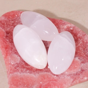 Può includere: Tre pietre ovali, bianche e rosa, poggiano su una base di pietra rosa strutturata. Le pietre hanno un aspetto liscio e lucido. La base ha una consistenza ruvida e naturale. Lo sfondo è beige neutro.
