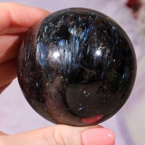 May include: A polished, dark sphere with a swirling pattern of blue and black. The gemstone ball has a reflective surface, catching light and creating a shimmering effect. The sphere is held in a hand, showcasing its size and detail.