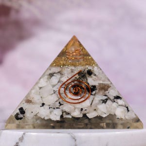 May include: A clear crystal pyramid with a copper wire spiral inside. The pyramid is filled with white and black crystals and gold glitter. It is sitting on a white marble base with a gold trim.