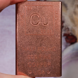 May include: A rectangular copper bar featuring the periodic table symbol Cu, atomic number 29, atomic mass 63.55, melting point 1085 degrees Celsius, and density 8.92 g/cc. The bar is marked HALF POUND, 999 FINE COPPER, and USA with two stars.