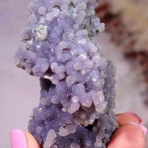 May include: A cluster of grape agate featuring numerous small, spherical, purple-coloured crystal formations. The mineral specimen displays a botryoidal habit with a translucent, crystalline texture across its surface, showcasing varying shades of light to deep violet throughout the entire structure.