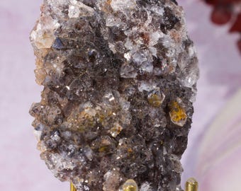 May include: A cluster of crystals in shades of gray, brown, and clear, mounted on a gold-colored metal stand. The crystals have a rough, textured surface and reflect light, creating a sparkling effect. The background is a soft, blurred pink and red.