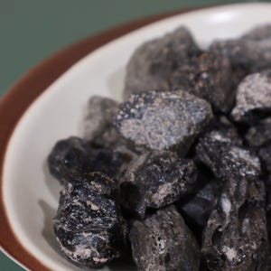 May include: A white bowl with a brown rim filled with dark grey, irregularly shaped rocks. The rocks have a rough texture and a slightly dusty appearance. The bowl sits on a green surface.