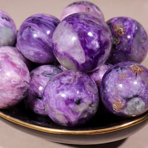 May include: A collection of polished charoite spheres in varying shades of purple and white, displayed in a dark bowl. The spheres have a swirling, marbled appearance, with some showing hints of black and brown.