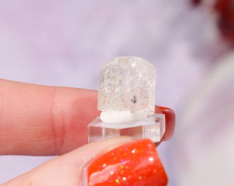 May include: A small, clear crystal sits atop a clear, square base. The crystal is held between fingers with bright orange glitter nail polish. The background is a soft, blurred purple.
