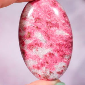 May include: An oval-shaped pink gemstone with white and darker pink veining. The polished stone is held between fingers, highlighting its smooth surface and natural colour variations. A close-up view shows the stone's unique patterns.