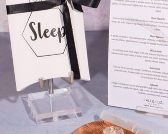 May include: A white gift box with a black ribbon and the word "Sleep" in a geometric design. A copper-colored dish holds several crystals, including rose quartz and amethyst. A card describes the crystals' properties.