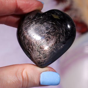 May include: A heart-shaped, polished stone with a dark, metallic appearance. The stone has swirling patterns and a reflective surface, with hints of silver and gold. The overall color is a deep black with iridescent highlights.
