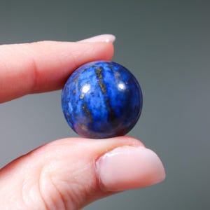 May include: A smooth, round, blue lapis lazuli sphere with gold flecks. The sphere is being held between two fingers.