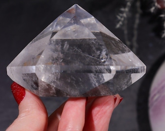 May include: A clear quartz crystal point with a diamond shape. The crystal is held in a hand with red nail polish. The crystal has many facets and is transparent, allowing light to pass through. The background is a dark gray.