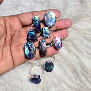 Puede incluir: Una colección de anillos de plata con conchas de abulón iridiscentes en varias formas, incluyendo ovaladas, rectangulares y de lágrima. Las conchas muestran una gama de colores, como azul, púrpura y verde. Los anillos se exhiben sobre una superficie blanca y esponjosa.