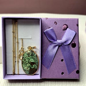 May include: A gold-toned chain necklace with an oval pendant featuring a mottled green and brown stone, presented in a light purple gift box with a matching purple ribbon bow on the lid. The necklace is displayed inside the open box.