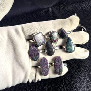 Peut inclure: Collection de bagues en pierres précieuses avec des montures en argent. Les bagues présentent des pierres de différentes formes, dont ovales, en goutte et rectangulaires. Les pierres affichent un mélange de couleurs, principalement violet foncé, vert et noir, avec un motif tacheté.