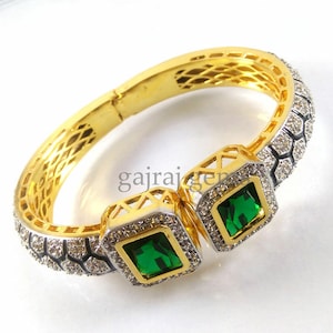May include: Gold bangle bracelet with two square green gemstones surrounded by clear stones. The bracelet has a textured design and is open on one side.